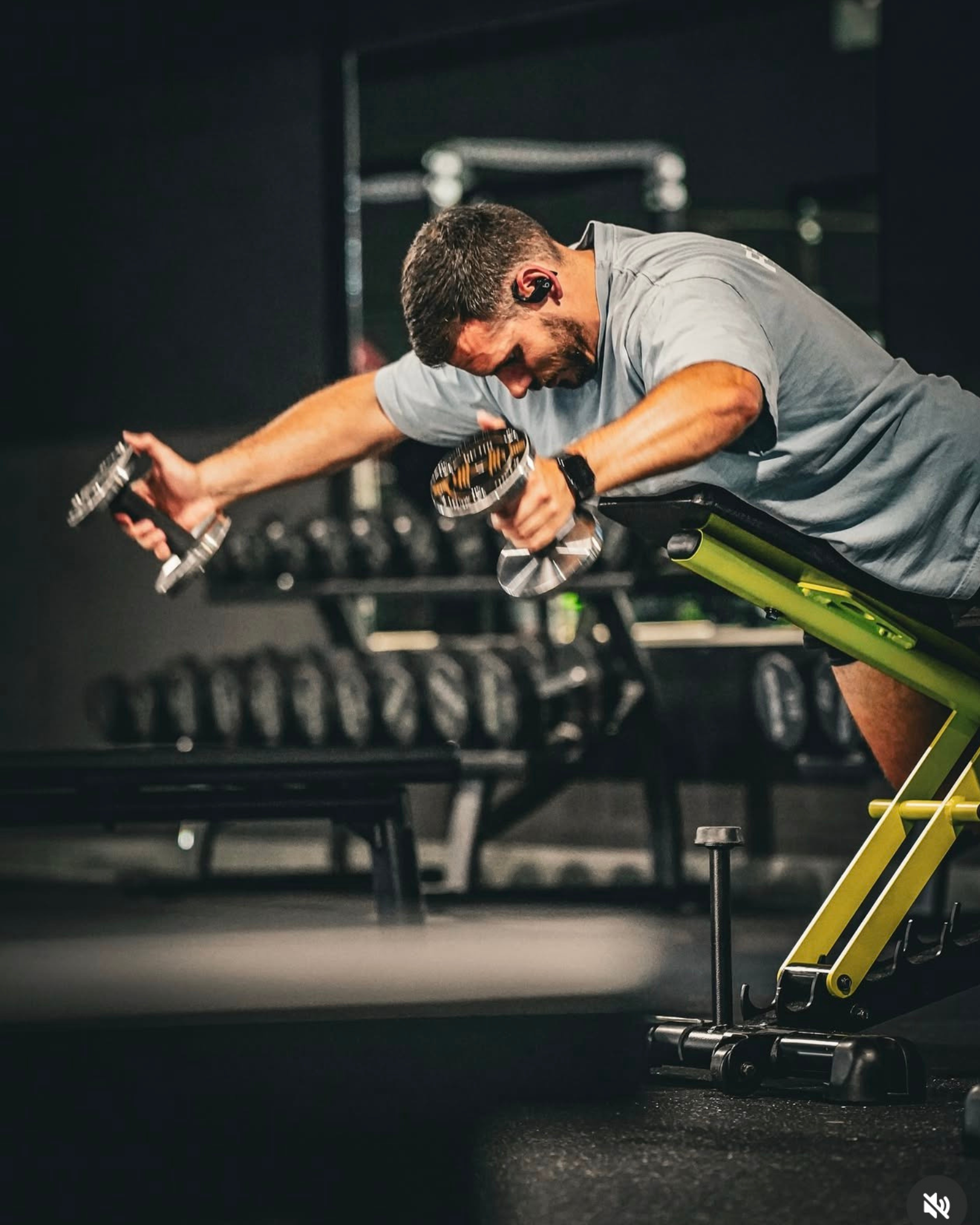 Man exercising on a sit-up bench at the gym, lifting dumbbells and wearing wireless earbuds.