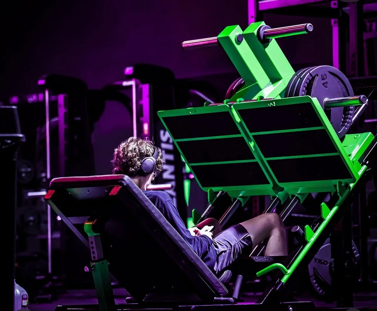 Person with curly hair wearing headphones lying on a workout bench in a gym with high-tech exercise equipment illuminated by purple and green lighting.