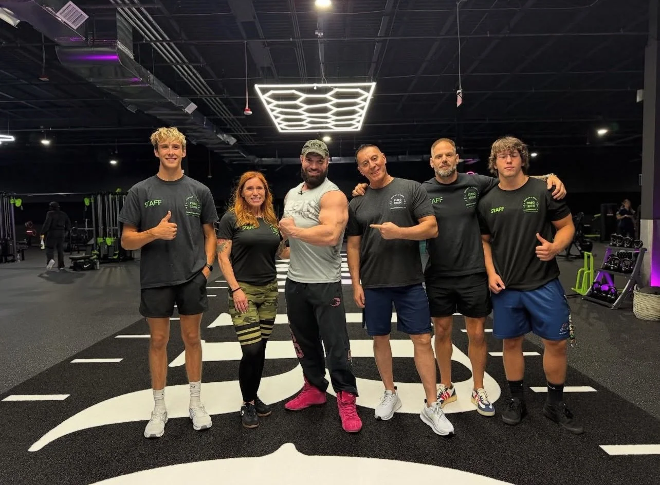 Group of six fitness trainers posing inside a gym, standing on a black and white logoed mat, with fitness equipment in the background.