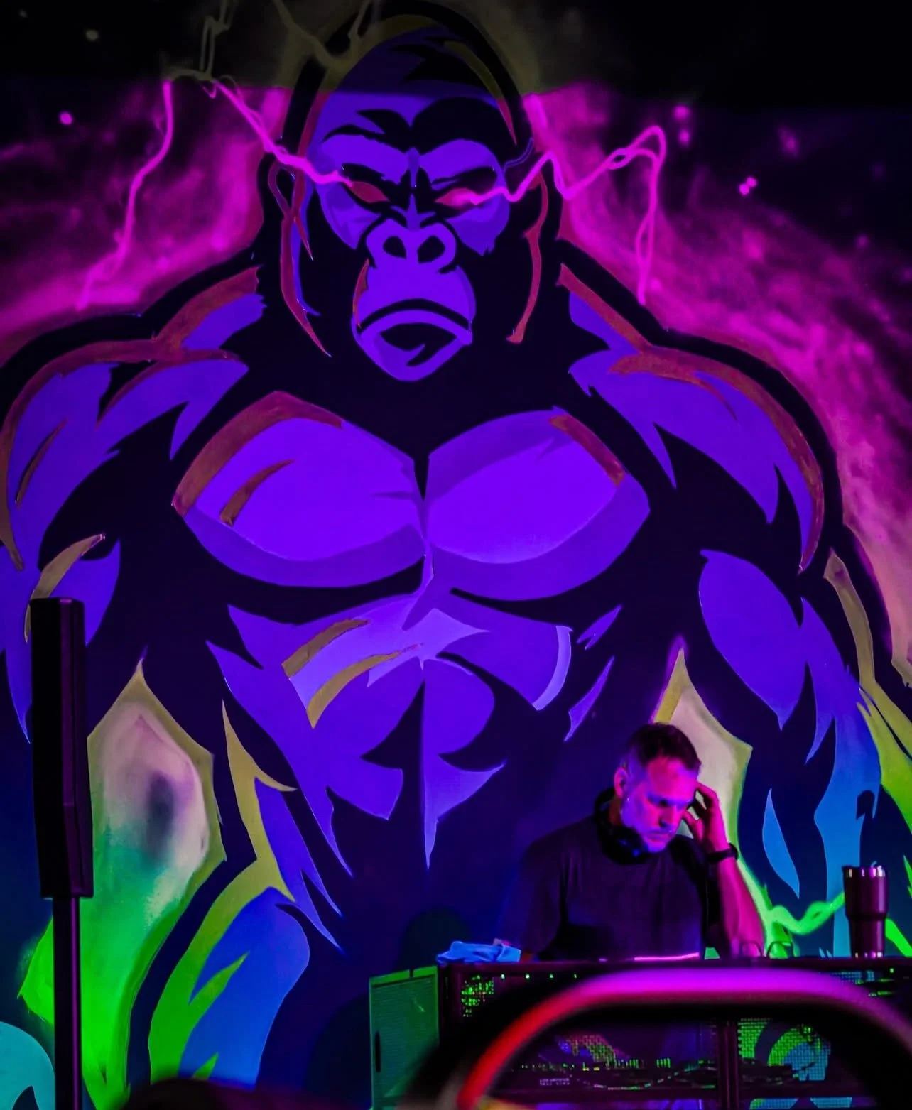 A DJ wearing a black shirt is performing with headphones behind a mixing console, in front of a large mural of a muscular gorilla with pink lightning effects, painted in glowing purple, pink, and green colors.