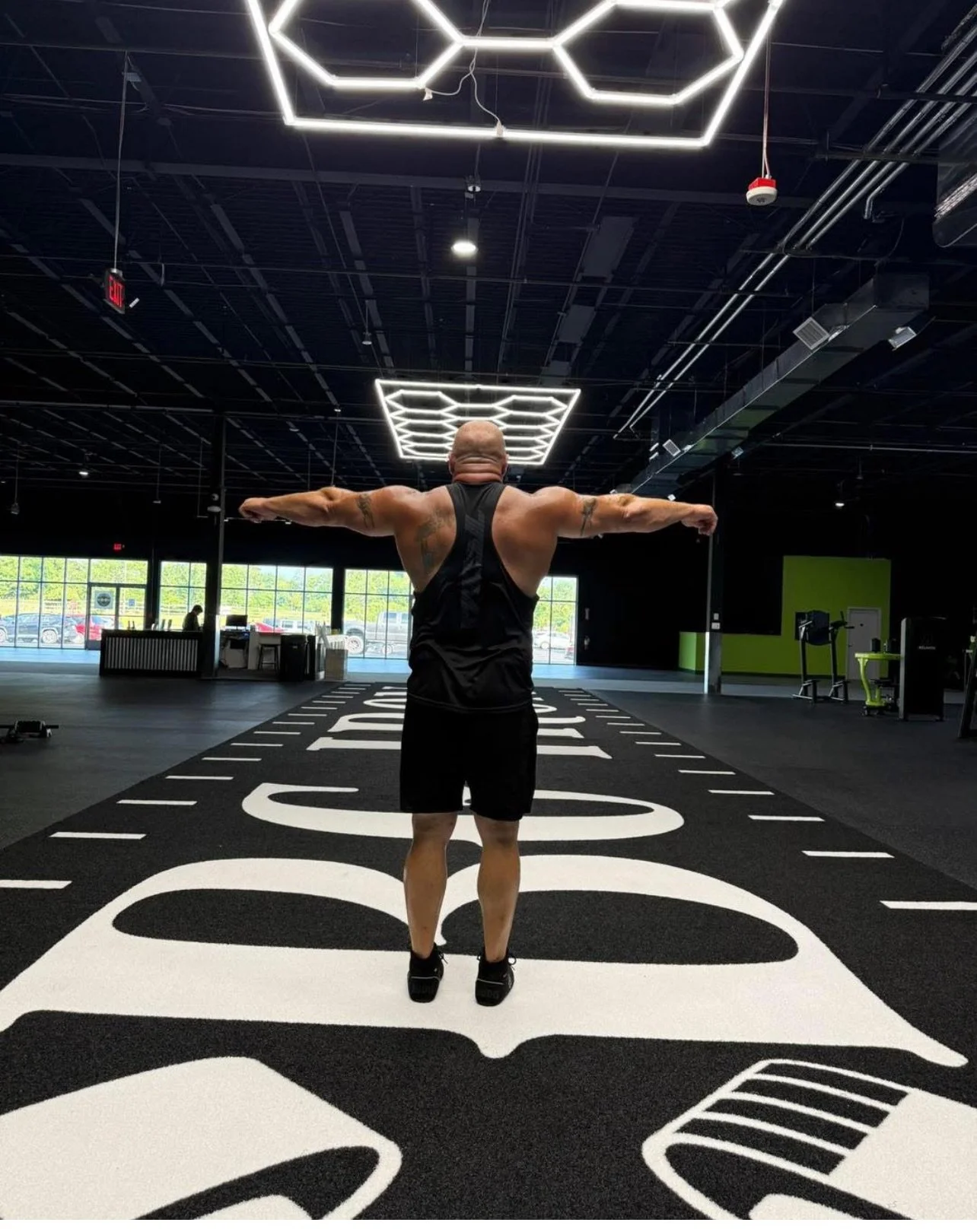 A man with a muscular build and tattoos on his arms is standing with arms outstretched inside a modern gym. The gym has black walls, a large window, and bright LED lighting fixtures on the ceiling. The floor features a large logo or design in black a