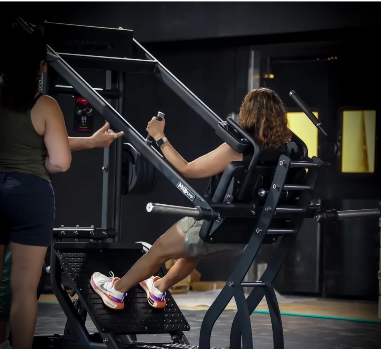 A woman using a leg press machine in a gym, assisted by another person, with dark walls and yellow lighted mirrors in the background.