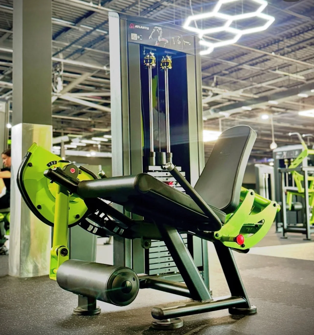 A green and black leg press exercise machine in a gym, with a weight stack and display, set against other gym equipment and a person in the background.