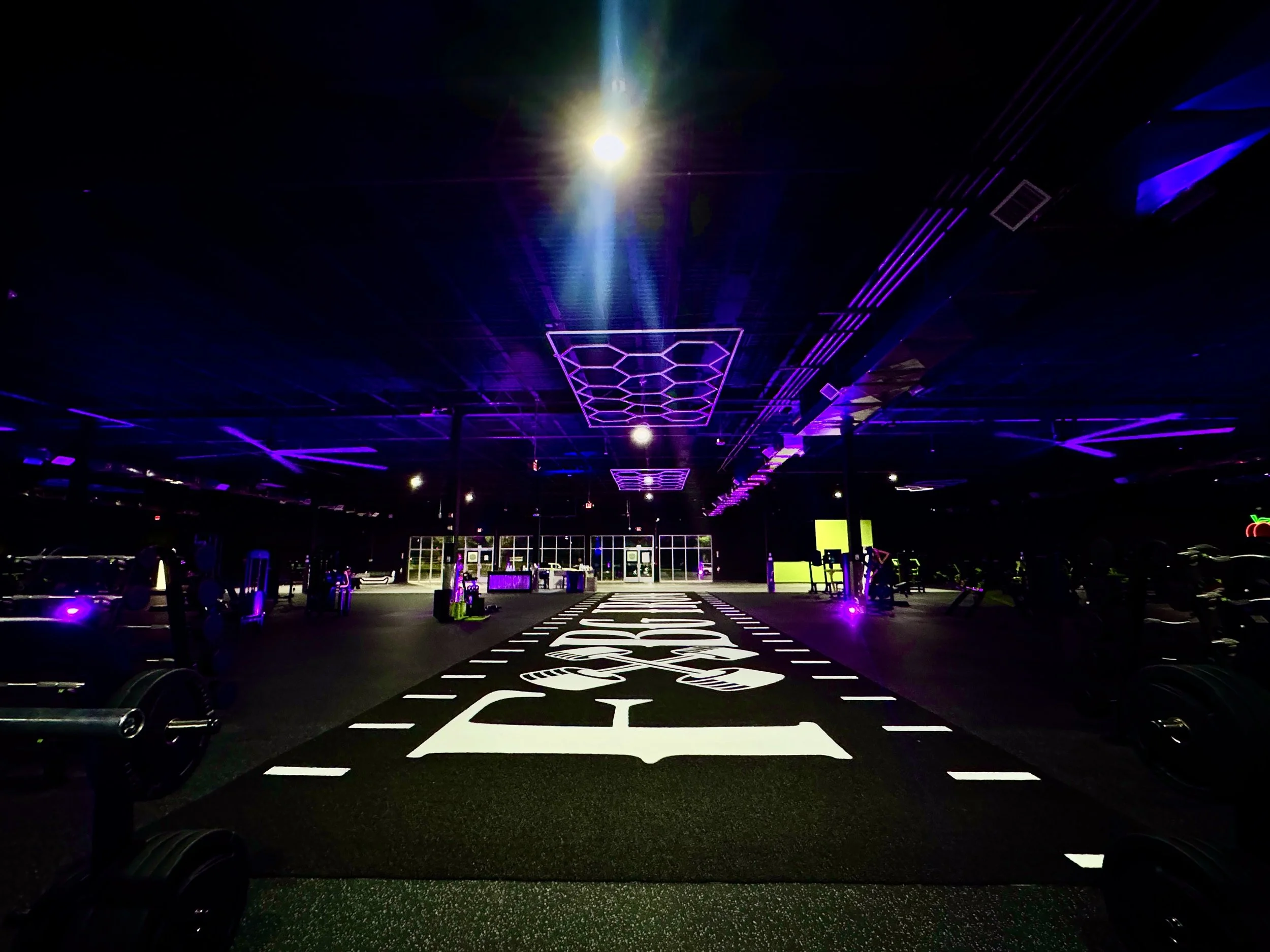 Indoor gym with purple and blue neon lighting, a black running track with white markings, and exercise equipment in the background.