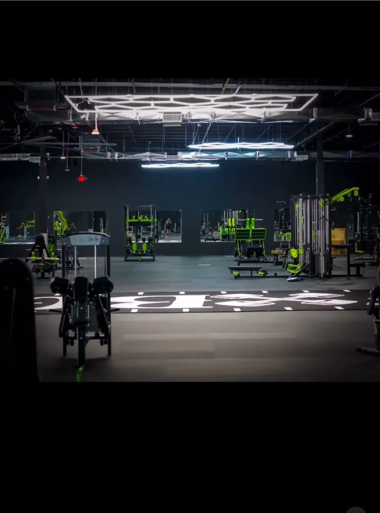 Empty gym with black walls, illuminated ceiling lights, and green and black workout equipment, including cardio machines, weight machines, and benches.