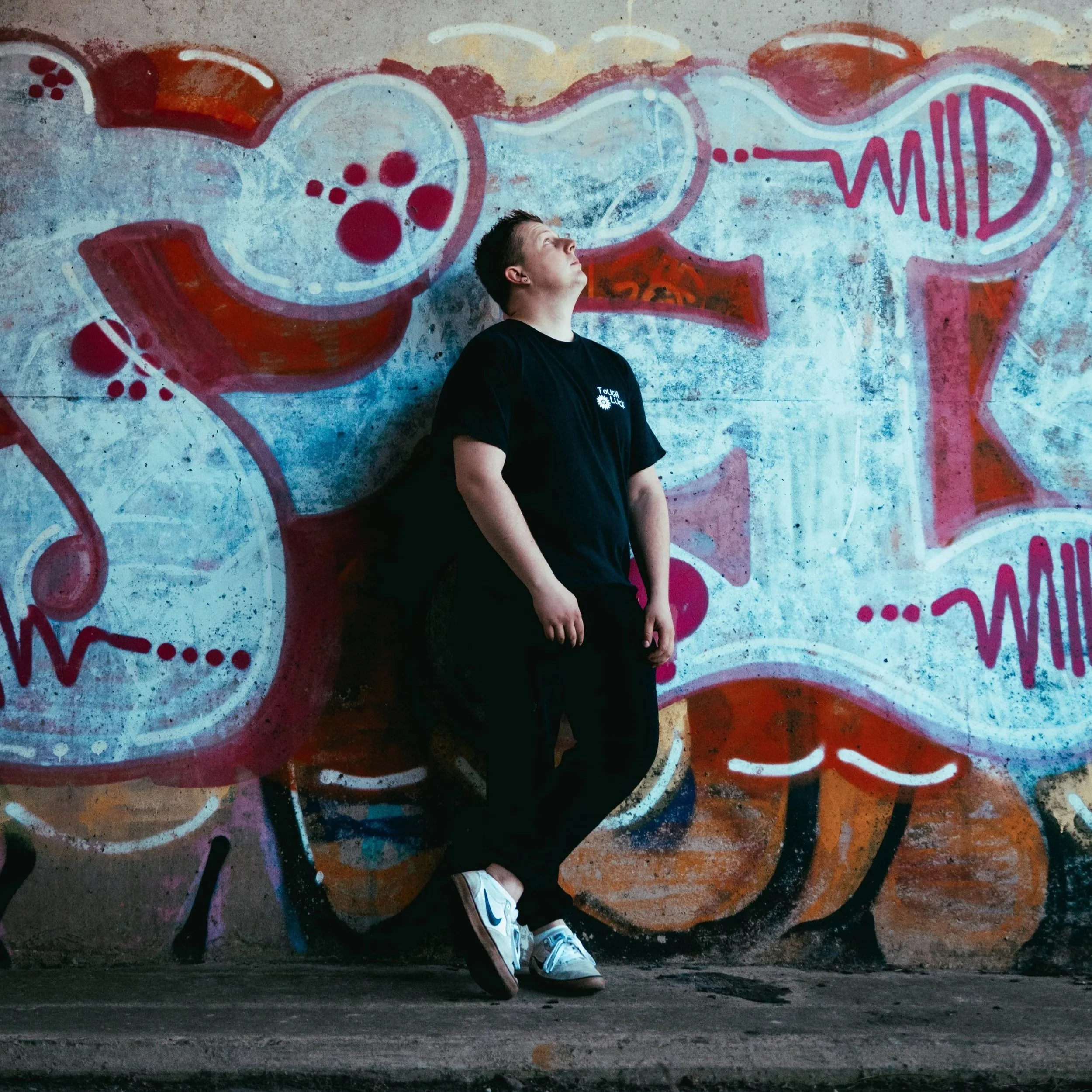 VIEK in a black shirt leaning against a graffiti-covered wall, looking upward.