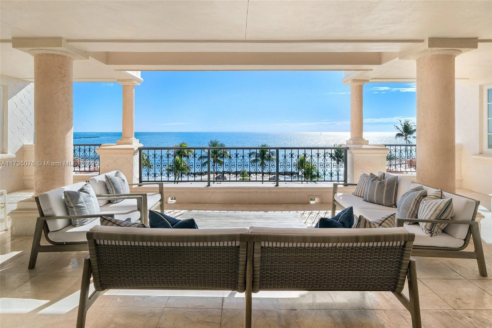 Luxury outdoor patio with white cushioned seating, overlooking a beach with palm trees and blue ocean under a clear sky.