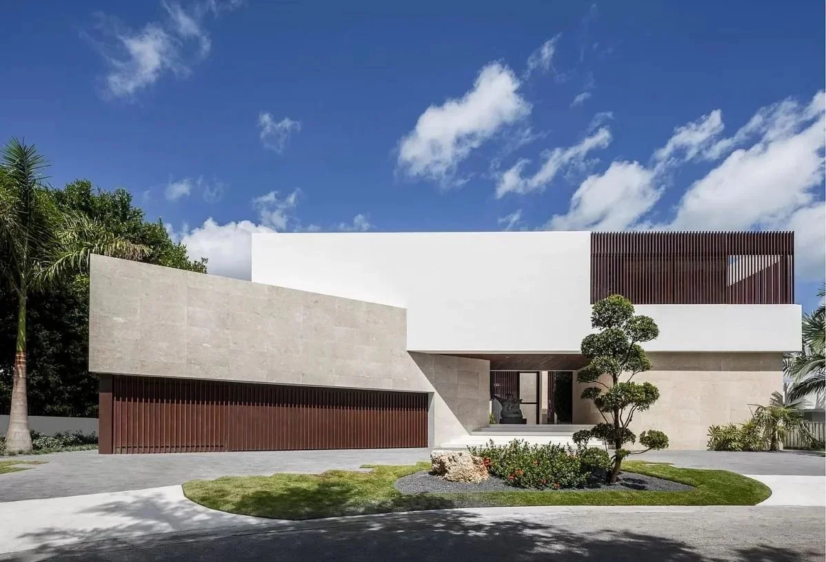 Modern residential house with white and beige walls, brown wooden accents, a small landscaped garden with a tree and rocks, and a paved driveway, under a clear blue sky with some clouds.