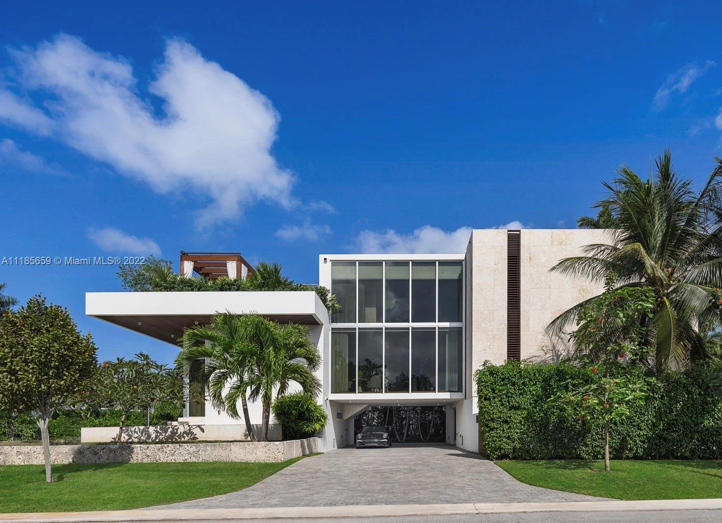 Modern two-story house with large glass windows, surrounded by palm trees and greenery, under a blue sky with clouds.