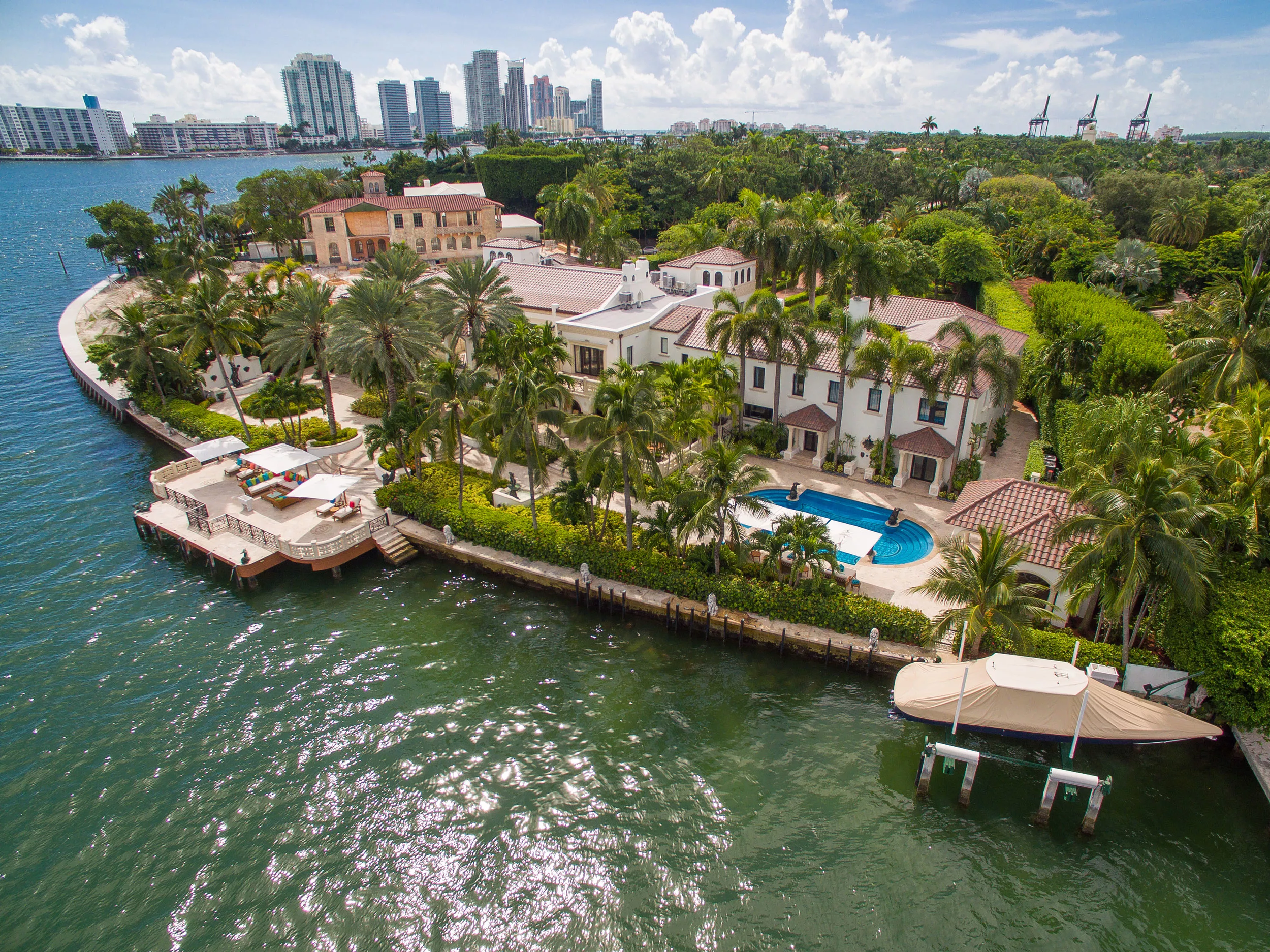 Luxury waterfront estate with a swimming pool, surrounded by palm trees, ocean, and city skyline in the background.