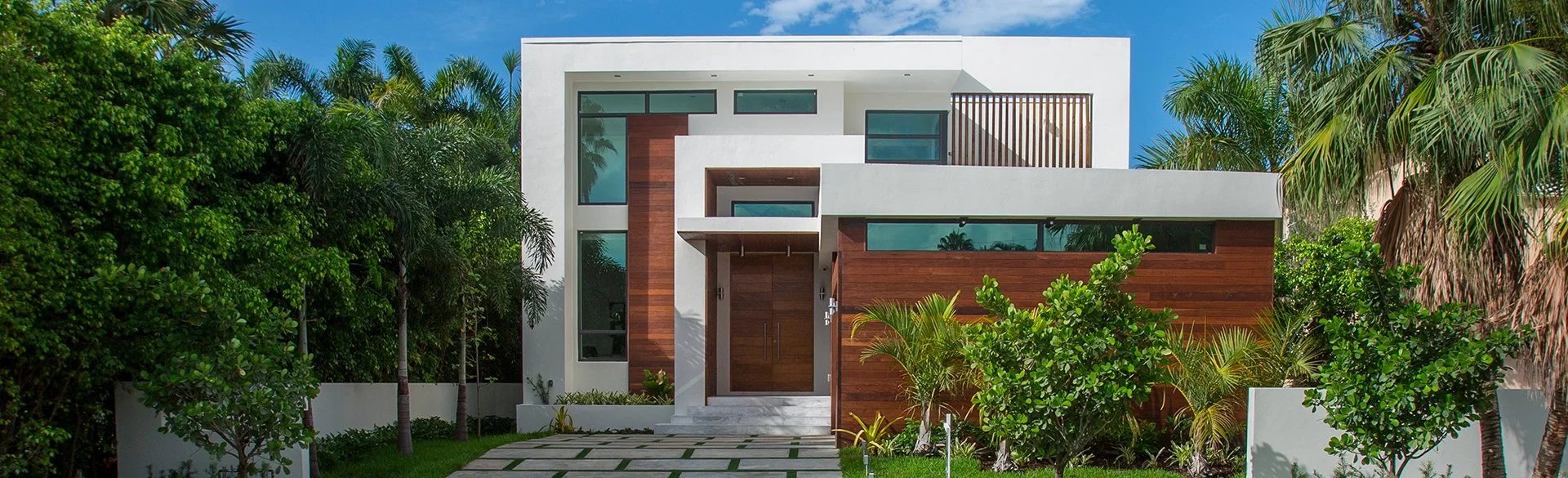 Modern two-story house with white and wood exterior, tall glass windows, surrounded by lush green palm trees and landscaping, under a blue sky.
