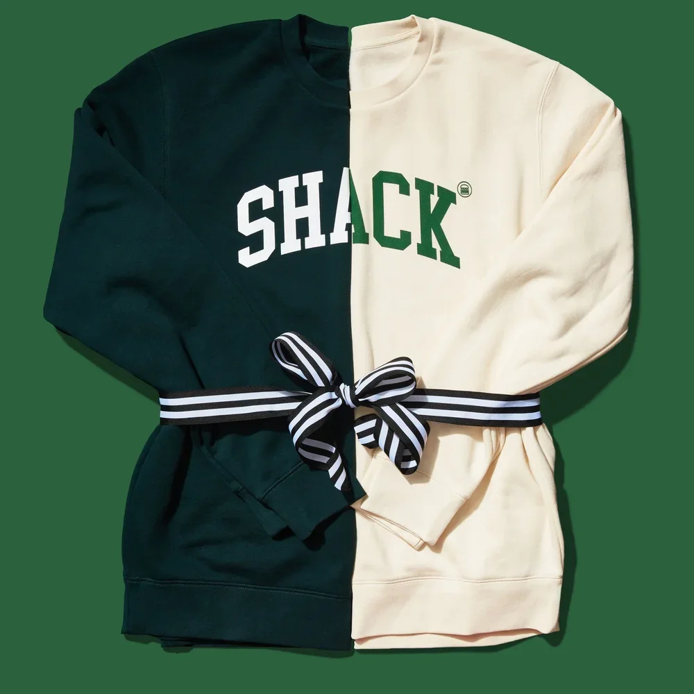 A split-colored sweatshirt, half green with 'SHACK' in white letters, half cream with 'SHACK' in green letters, with a striped black and white ribbon around it, on a green background.