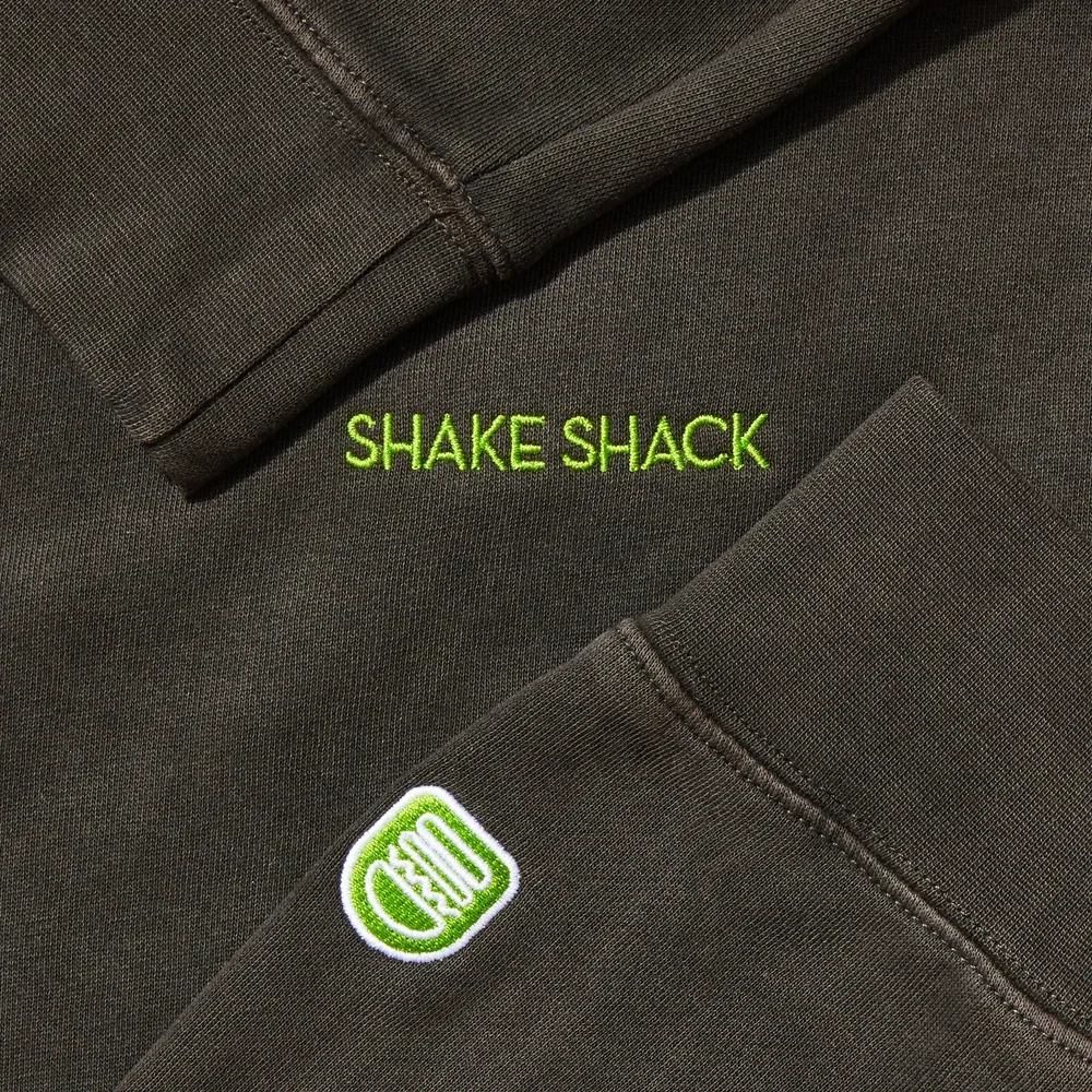 Close-up of a black Shake Shack uniform with green embroidered text that says "SHAKE SHACK" and a green cactus logo patch.