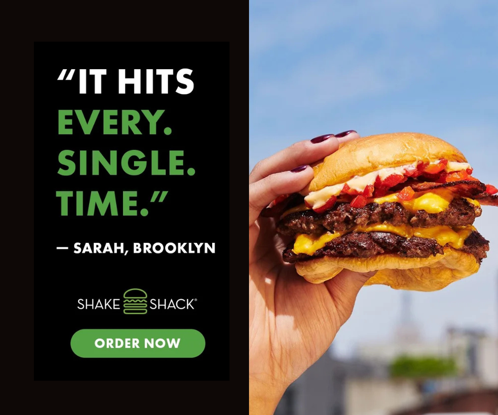 A person holding a large burger with three beef patties, melted cheese, bacon, and toppings, outdoors with a blue sky.