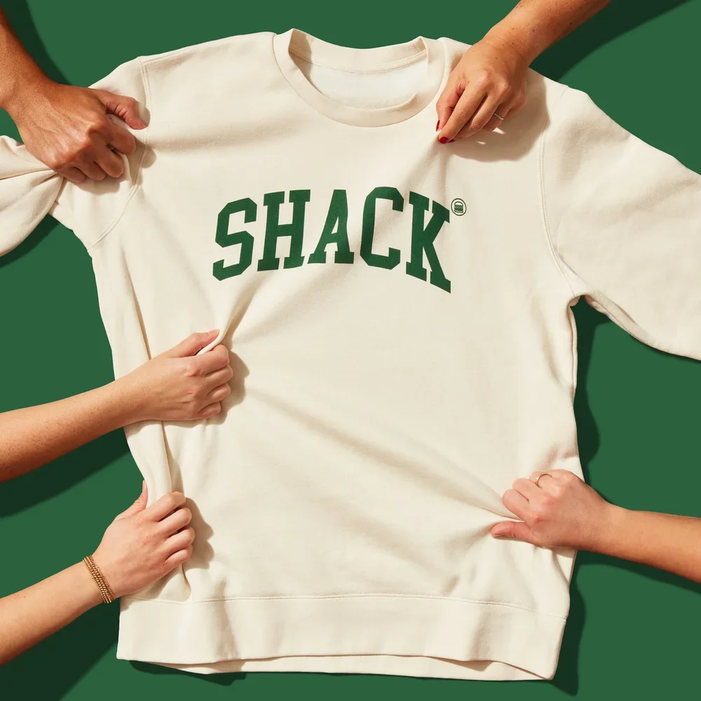 Four people holding a white sweatshirt with the word "SHACK" in large green letters.