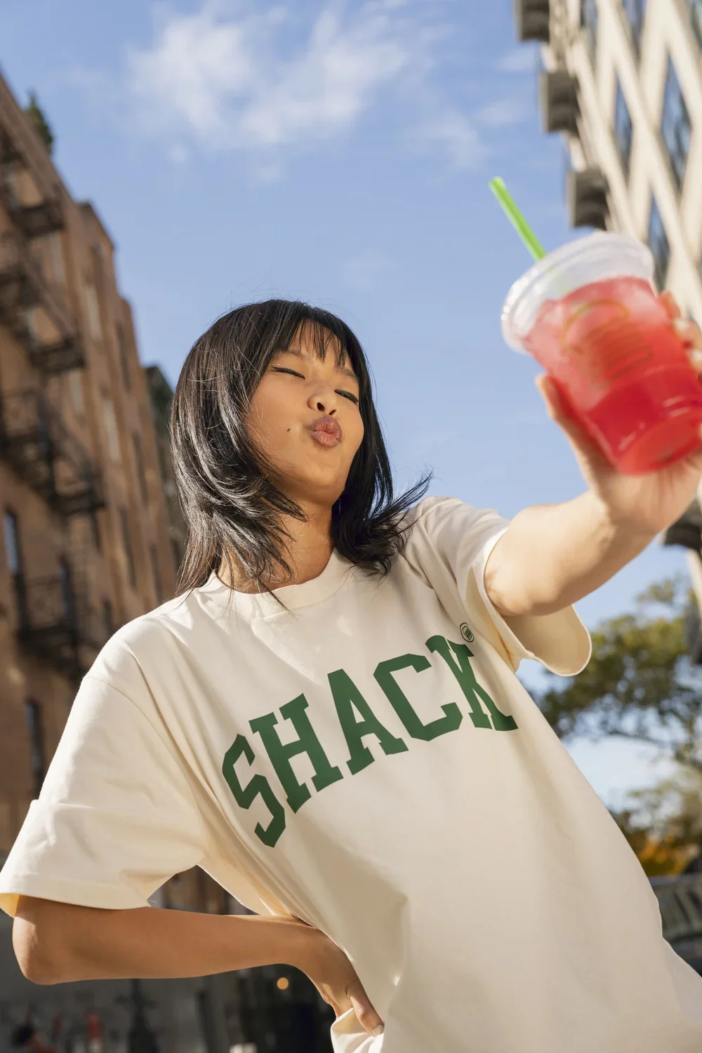 Young woman outdoors taking a selfie with a colorful drink, wearing a beige t-shirt with the word 'SHACK' printed on it, making a playful pouty face, with buildings and blue sky in the background.