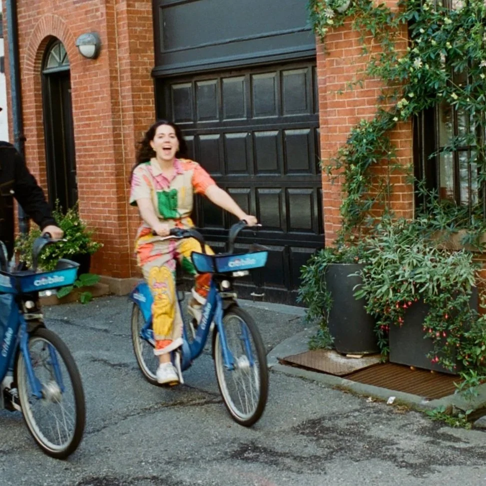 A woman riding a blue bicycle, smiling and looking excited, with a brick building and green plants in the background.