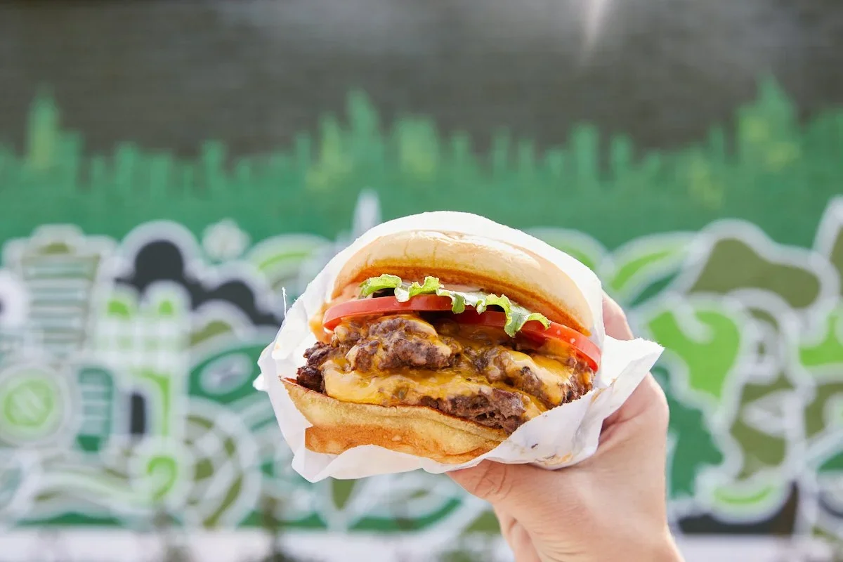 Hand holding a cheeseburger with lettuce, tomato, and cheese in front of a green graffiti wall.