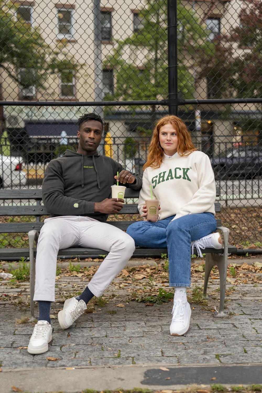 A man and woman sit on a park bench, holding drinks, in front of a chain-link fence with trees and apartment buildings in the background. The man wears a dark hoodie and white pants, and the woman wears a white sweatshirt and blue jeans.