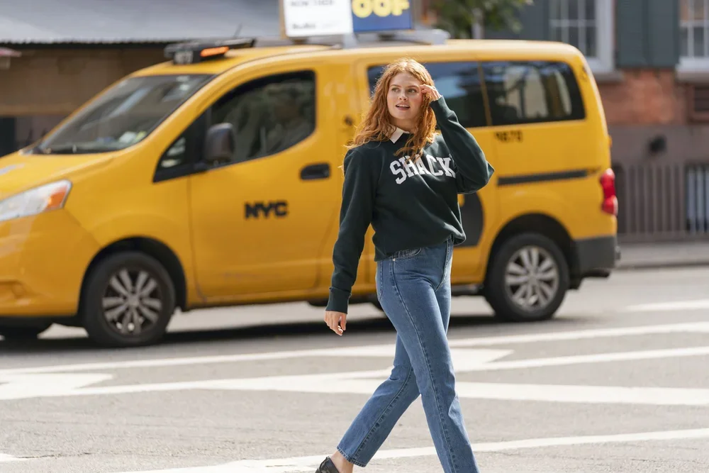 A young woman with curly red hair walking across a crosswalk in front of a yellow NYC taxi cab.