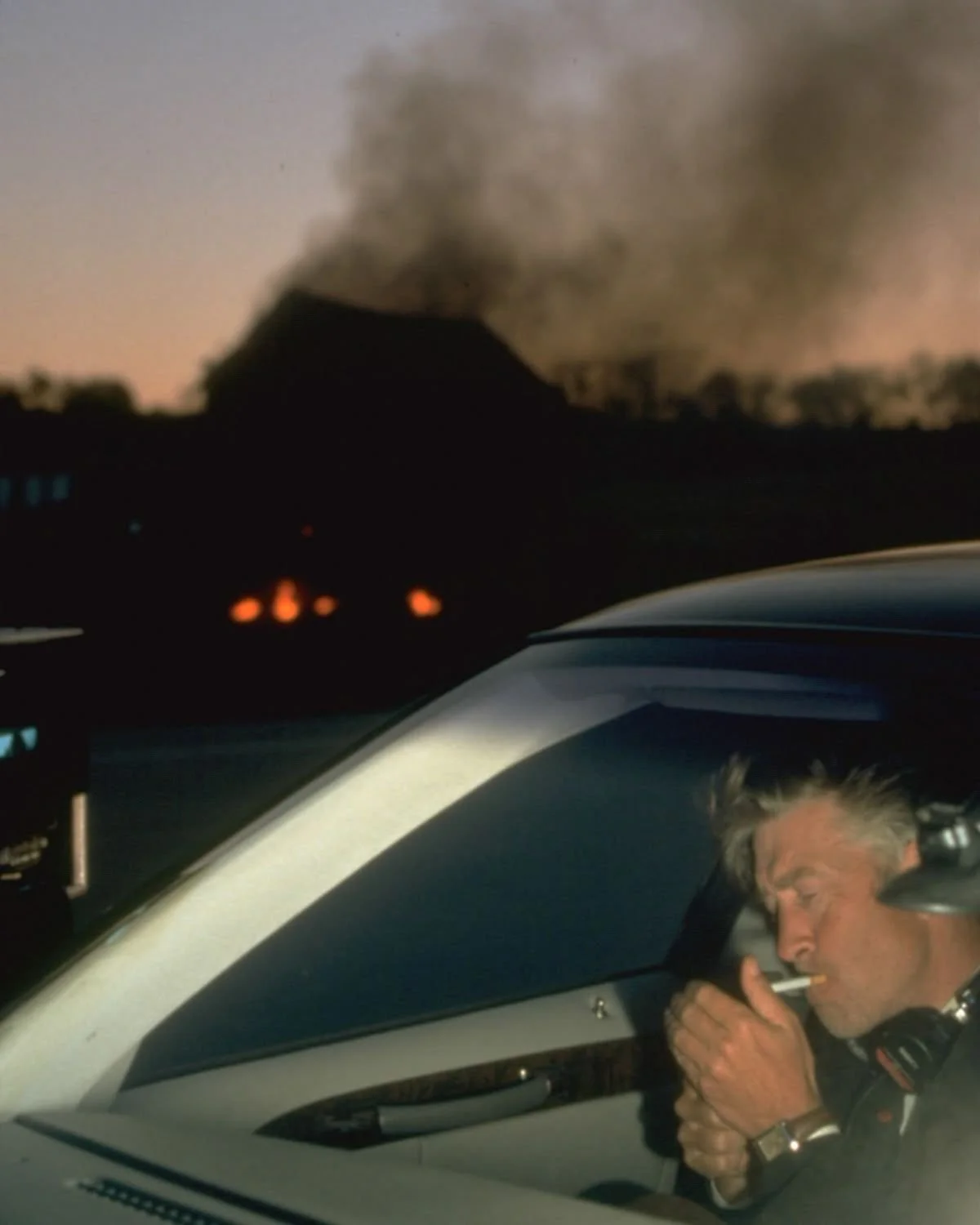 A man in a police uniform is sitting inside a police helicopter, lighting a cigarette, with a dark, smoky volcano in the background at dusk or dawn.