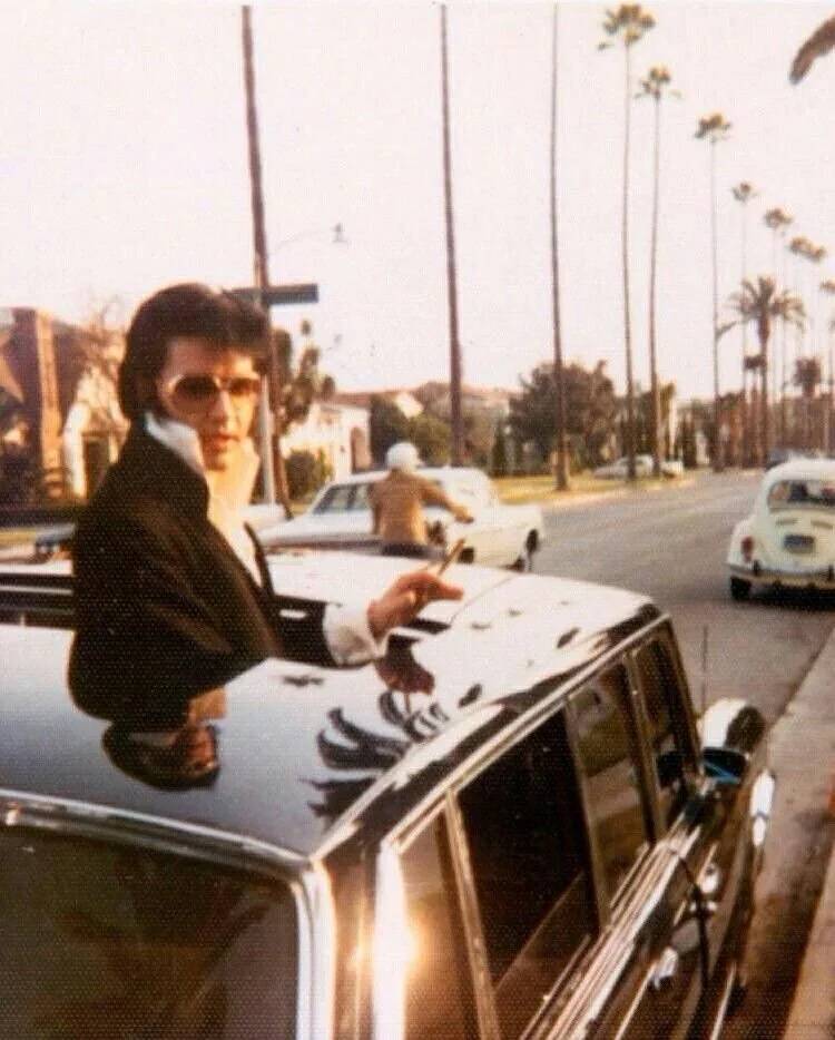 Woman with sunglasses standing through sunroof of black vintage car on a sunny street lined with palm trees and retro-style buildings.