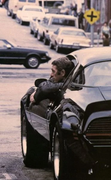 Man leaning out of a black sports car on a city street, with parked cars and a yellow pedestrian crossing sign in the background.