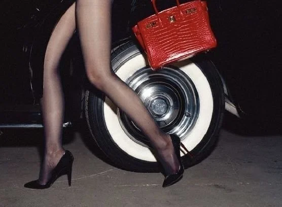 A woman wearing black high heels and sheer black stockings, with a red handbag, is standing next to a vintage car with white-wall tires.