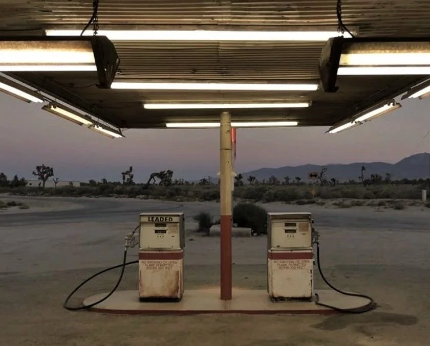 Empty gas station in a desert at dusk with two vintage gas pumps under a metal roof shelter, surrounded by sparse desert vegetation and mountains in the distance.
