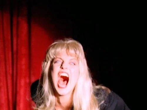A woman with blonde hair expressing anger or frustration, with her mouth open wide as if yelling, against a dark background with red curtains.