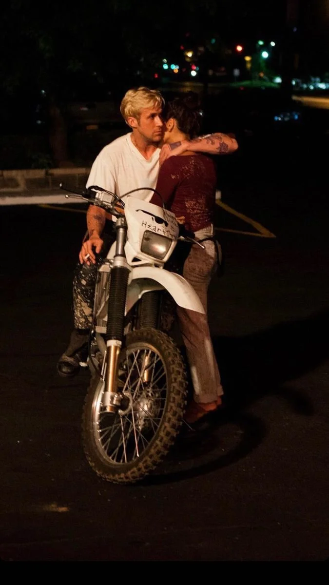 Two people, a man with blonde hair and a woman with short dark hair, are standing close together at night. The man is sitting on a white dirt bike, and the woman is hugging him while facing him. The background shows a dark street with blurred colorfu
