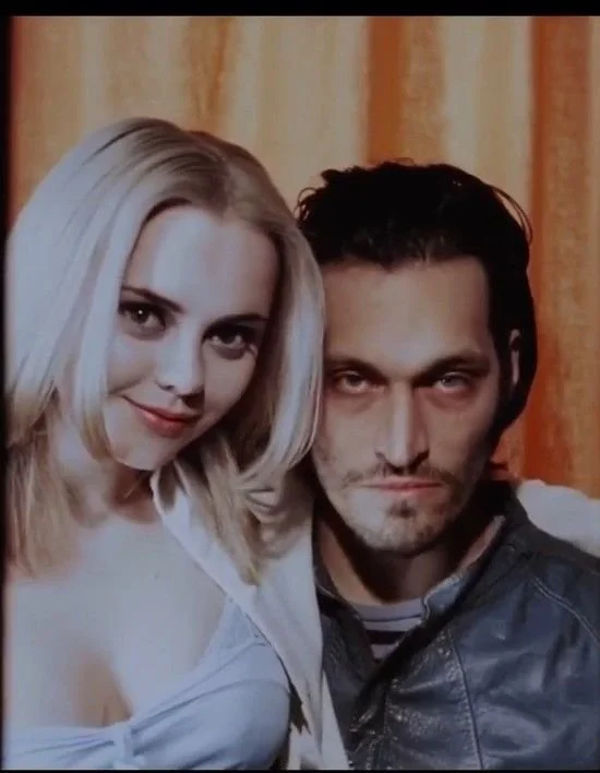 Photo of a young blonde woman and a man with dark hair, both looking at the camera, standing close together with a background of yellow and orange curtains.