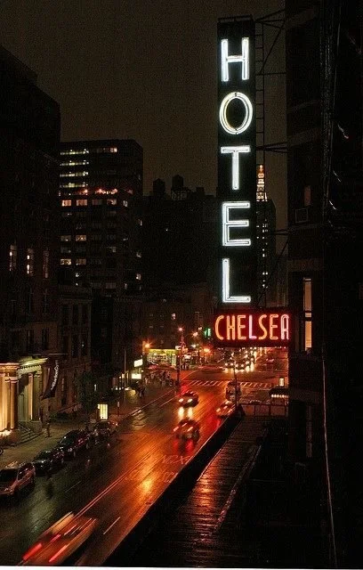 Nighttime city street view with illuminated hotel sign that reads 'HOTEL' vertically and 'CHELSEA' horizontally, with wet road reflecting lights and cars driving by.