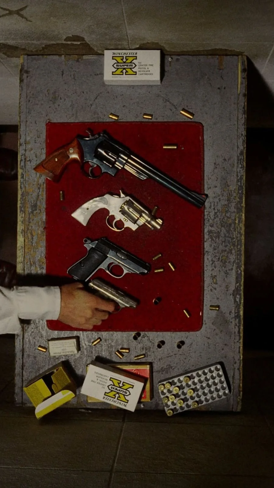 Three revolvers and one semi-automatic pistol on a red mat with handgun ammunition scattered around. A person is loading a magazine into the semi-automatic pistol. There are boxes of Winchester ammunition on the table.