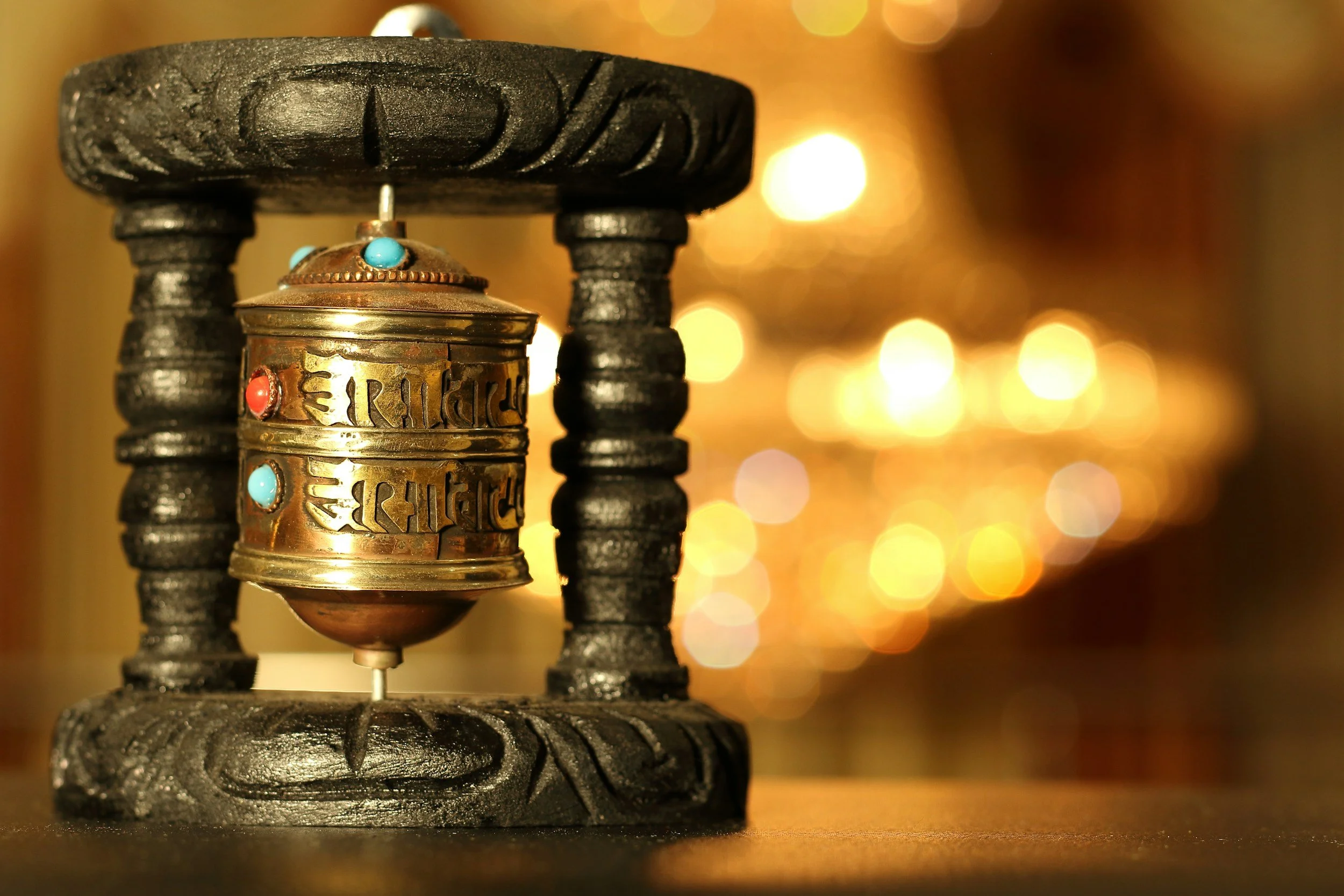 Close-up of a Tibetan prayer wheel with colorful stones, mounted on a wooden stand, with blurred warm lights in the background.