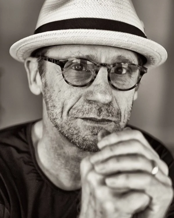 A black and white close-up portrait of a man wearing a white fedora hat with a black band and dark glasses. He has a serious expression, facial stubble, and is resting his chin on clasped hands.