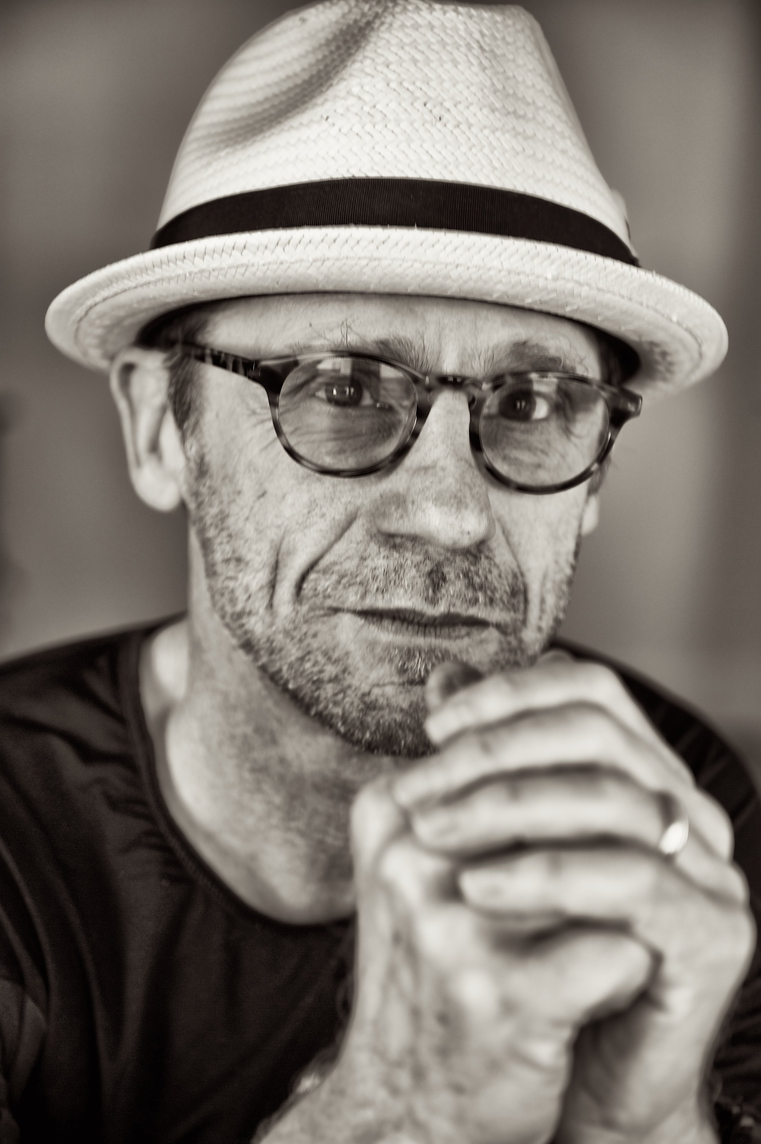 A man wearing a straw fedora, glasses, and a black shirt, with his hands clasped near his face, looking contemplative.