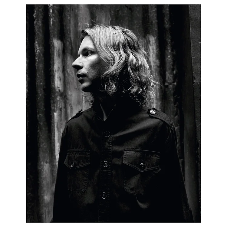 A black-and-white photo of a person with shoulder-length wavy hair, wearing a dark jacket, standing in front of a textured wooden wall and looking to the left.
