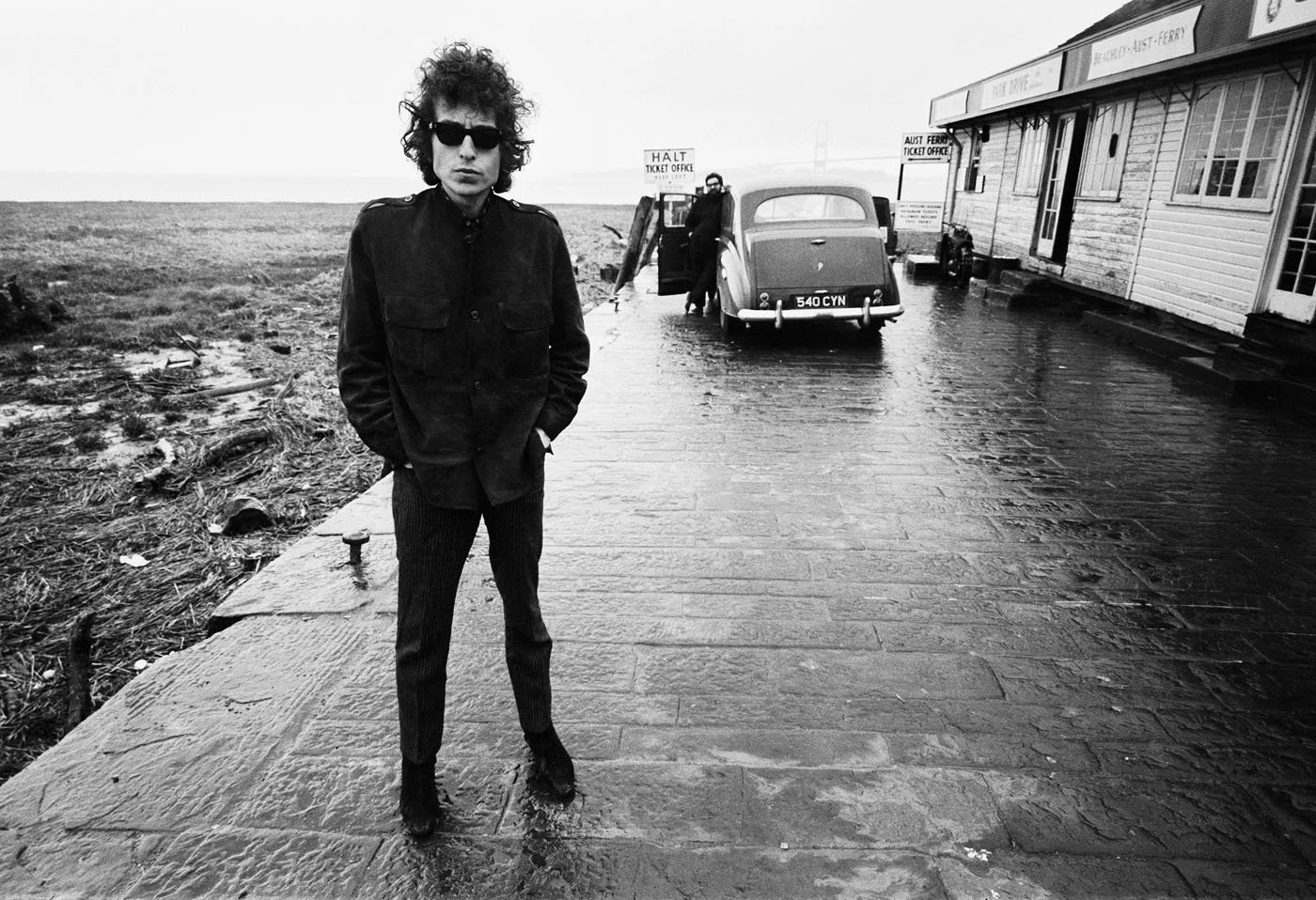 A man with curly hair wearing sunglasses, a dark jacket, and striped pants standing on a wet sidewalk near a rustic building with a sign that says 'Aust Ferry Ticket Office.' A vintage car is parked nearby, and the background features a foggy landscape and a bridge.