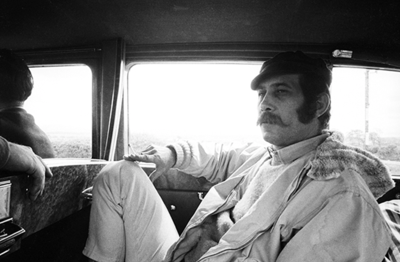 Black-and-white photo of a man with a mustache and sideburns sitting in the backseat of a car, looking out the window with a serious expression.