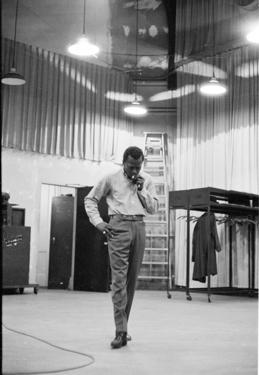 A man in dress shoes, slacks, and a button-up shirt standing on a stage with one hand in his pocket and looking down, holding a cigarette in his other hand. The room has stage curtains, hanging lights, and a ladder in the background.