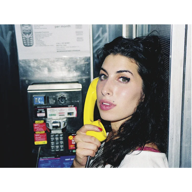 Woman with dark hair and red nails using a yellow payphone in front of a payphone booth.