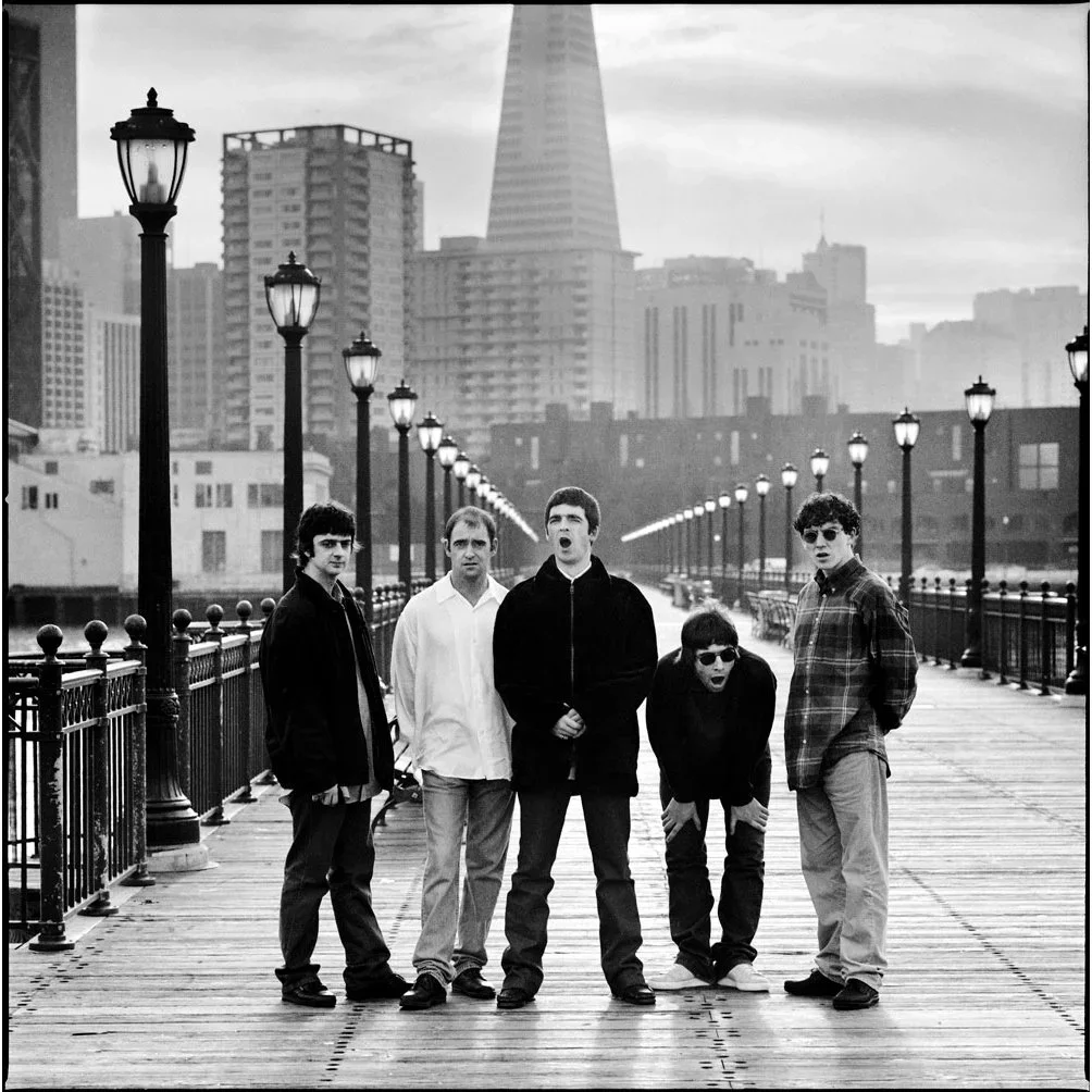 Five young men standing on a wooden pier with city skyscrapers in the background, some with open mouths as if singing or talking, in black and white.