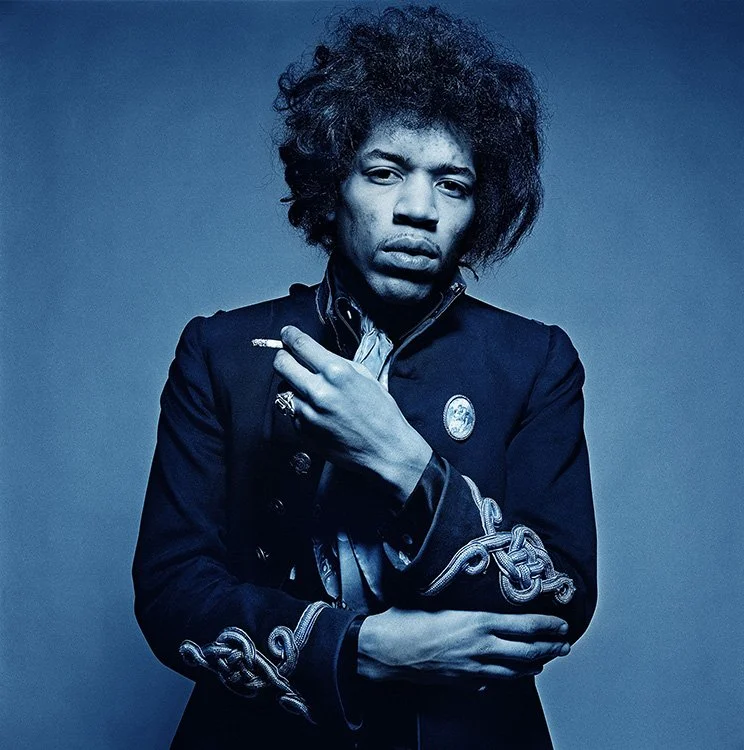 A young man with curly hair and a serious expression, wearing a dark jacket with decorative embroidery, holding a cigarette in one hand, against a plain blue background.