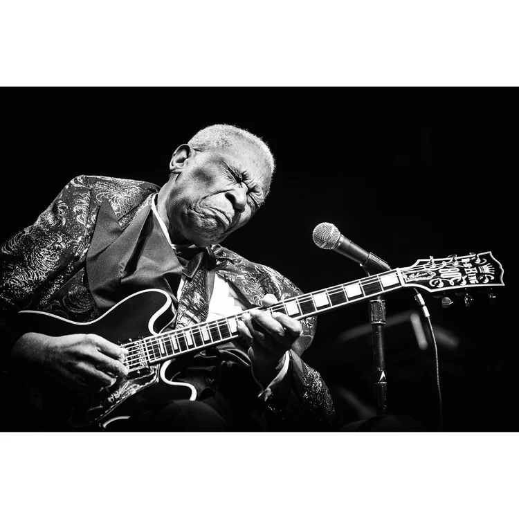 Black and white photo of an elderly man playing an electric guitar, eyes closed, with a microphone nearby, wearing a patterned jacket and a bow tie, on stage.