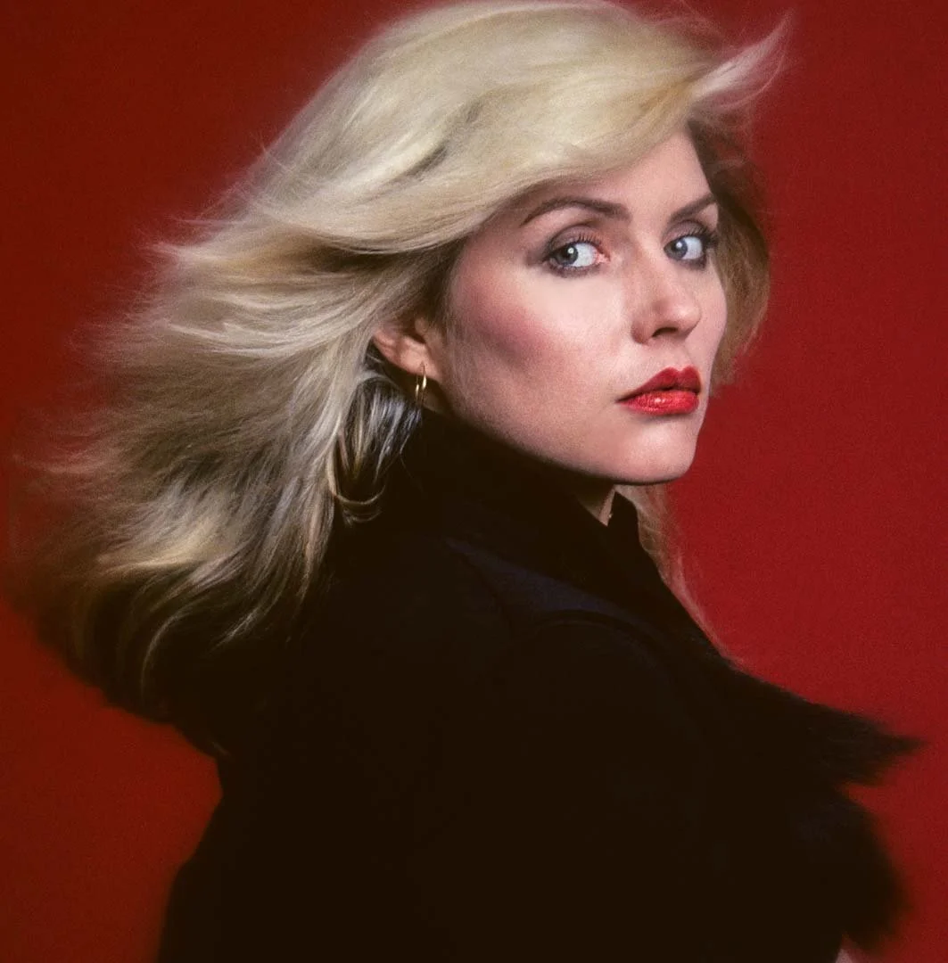 A woman with blonde, windswept hair, fair skin, red lipstick, wearing a black top and gold hoop earrings, posed against a red background.