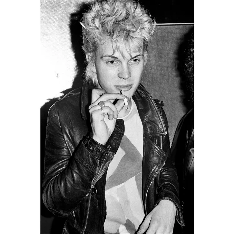 Black and white photo of a young man with curly, platinum blonde hair, wearing a leather jacket and a graphic t-shirt, posing with his hand near his chin.