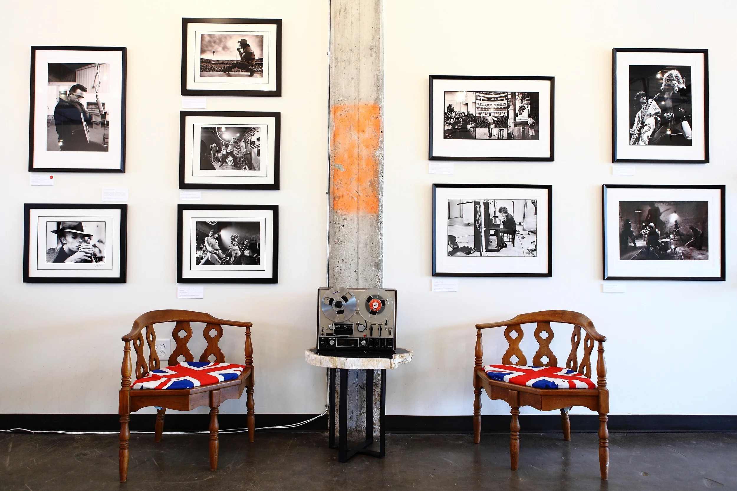 An art gallery with black and white photographs displayed in black frames on a white wall, two wooden chairs with British flag cushion covers, and a vintage reel-to-reel tape recorder on a small table in front of a concrete pillar.