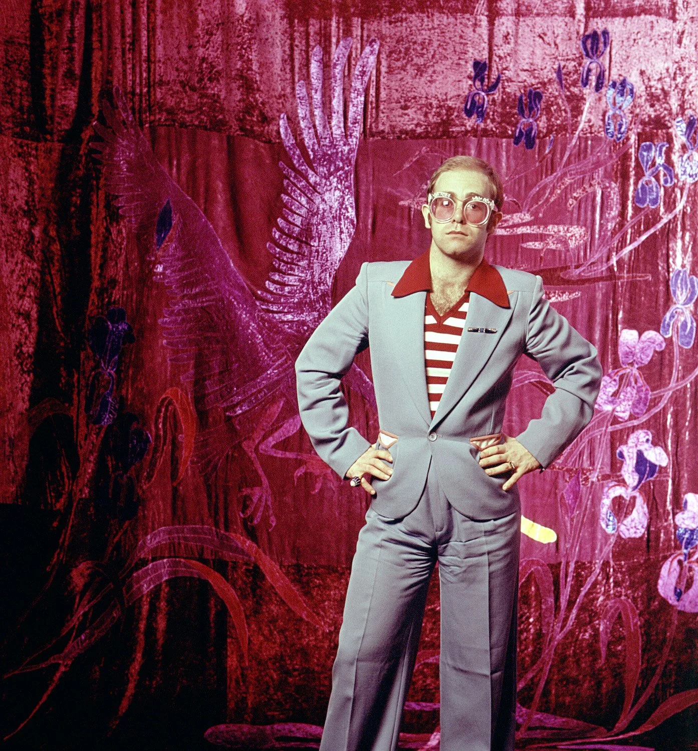 Man in gray suit with red collar shirt and striped shirt standing in front of a red curtain with purple flowers and large pink bird artwork.