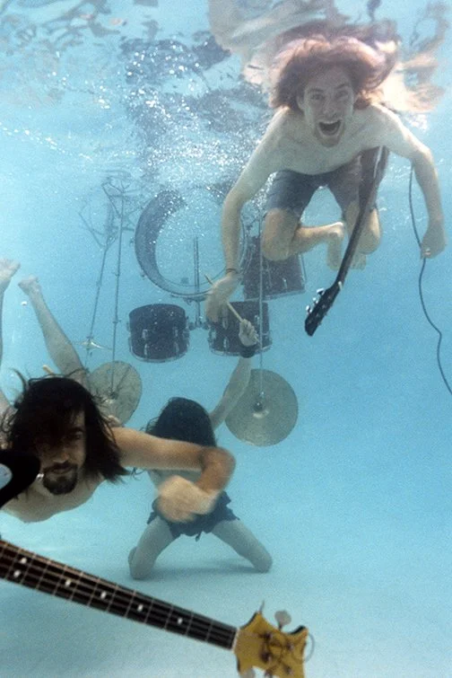 Three people underwater in a pool, with one person holding a guitar, a person holding a microphone, and a person in front of a camera or equipment.